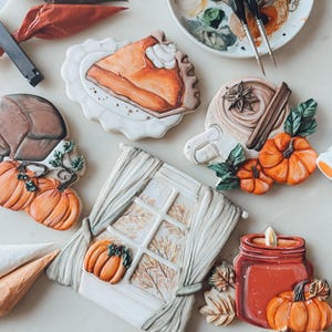 Könnte beinhalten: Eine Sammlung von Herbst-Zuckerplätzchen, die mit Herbstfarben und -mustern verziert sind. Die Plätzchen beinhalten einen Kürbiskuchen, ein Fenster mit Vorhängen, eine Tasse Kaffee mit Zimt, einen Kürbis und ein Kerzengefäß.