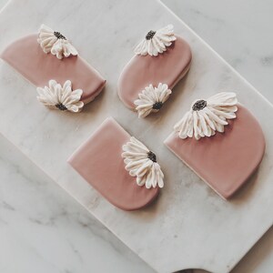 Pode incluir: Cinco biscoitos de a&ccedil;&uacute;car cor-de-rosa decorados com glac&ecirc; branco e flores brancas. Os biscoitos est&atilde;o dispostos sobre uma superf&iacute;cie de m&aacute;rmore branco.