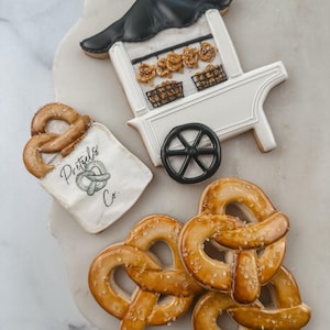 Puede incluir: Imagen de galletas con forma de pretzel. Incluye un carrito de pretzels blanco con techo negro, una bolsa con el texto "Pretzels Co." y varios pretzels individuales. Los pretzels son de color marrón dorado y están espolvoreados con sal.