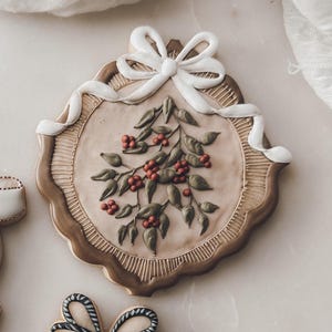 May include: A round, brown, gingerbread cookie decorated with a white bow and a green holly sprig with red berries.
