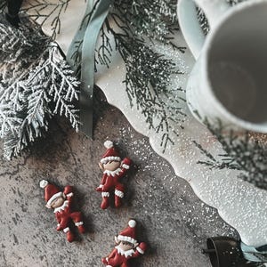 May include: A festive Christmas scene featuring three small elf figurines in red and white outfits. A white mug, a decorative plate with a snowy design, and sprigs of evergreen complete the holiday theme. The scene is set on a textured gray surface.