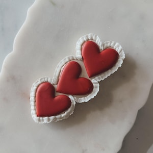 May include: Three red heart-shaped cookies with white ruffled edges. The cookies are arranged in a cluster on a white marble surface. The hearts are a vibrant red color, and the ruffled edges add a delicate touch.