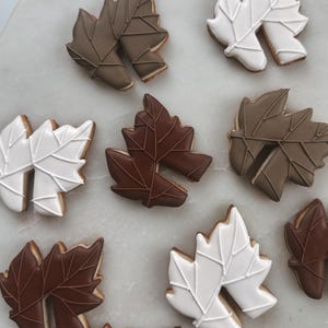 May include: An overhead shot displays several leaf-shaped cookies. The cookies are decorated with white, brown, and taupe icing, with detailed vein patterns. The cookies are arranged on a white surface, suggesting a fall or autumn theme.