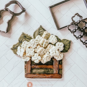 Puede incluir: Una galleta de flor de hortensia blanca en una maceta de madera. Las galletas están decoradas con glaseado real y están listas para disfrutar. Lolano Cookie Cutters © 2023.