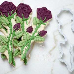 May include: A set of six purple and green rose cookie cutters with a single cutter in the foreground. The cutters are made of white plastic and are designed to create detailed rose cookies.