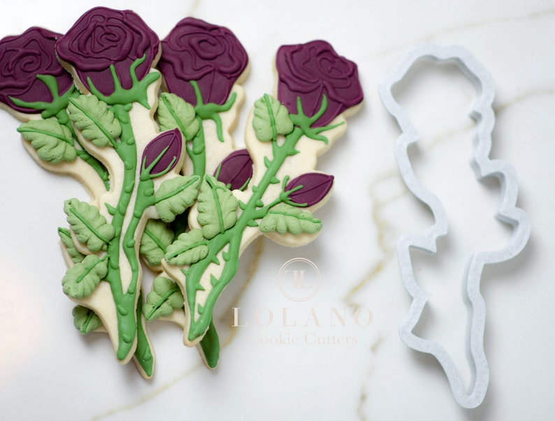 May include: A set of six purple and green rose cookie cutters with a single cutter in the foreground. The cutters are made of white plastic and are designed to create detailed rose cookies.
