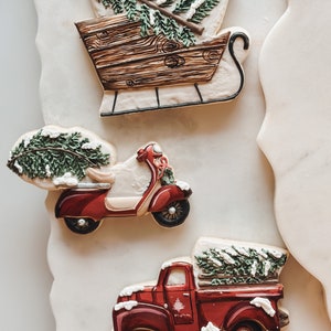 Könnte beinhalten: Drei weihnachtliche Zuckerkekse, verziert mit königlichem Zuckerguss. Ein roter Lastwagen, ein roter Roller und ein Holzschlitten, jeder mit einem grünen Weihnachtsbaum beladen.
