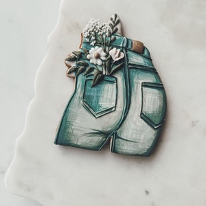 K&ouml;nnte beinhalten: Ein dekorierter Zuckerkeks in Form von Jeansshorts mit einem Blumenarrangement in der Ges&auml;&szlig;tasche. Der Keks ist hellblau mit wei&szlig;en Schattierungen und braunen Akzenten. Die Blumen sind wei&szlig;, rosa und gr&uuml;n.