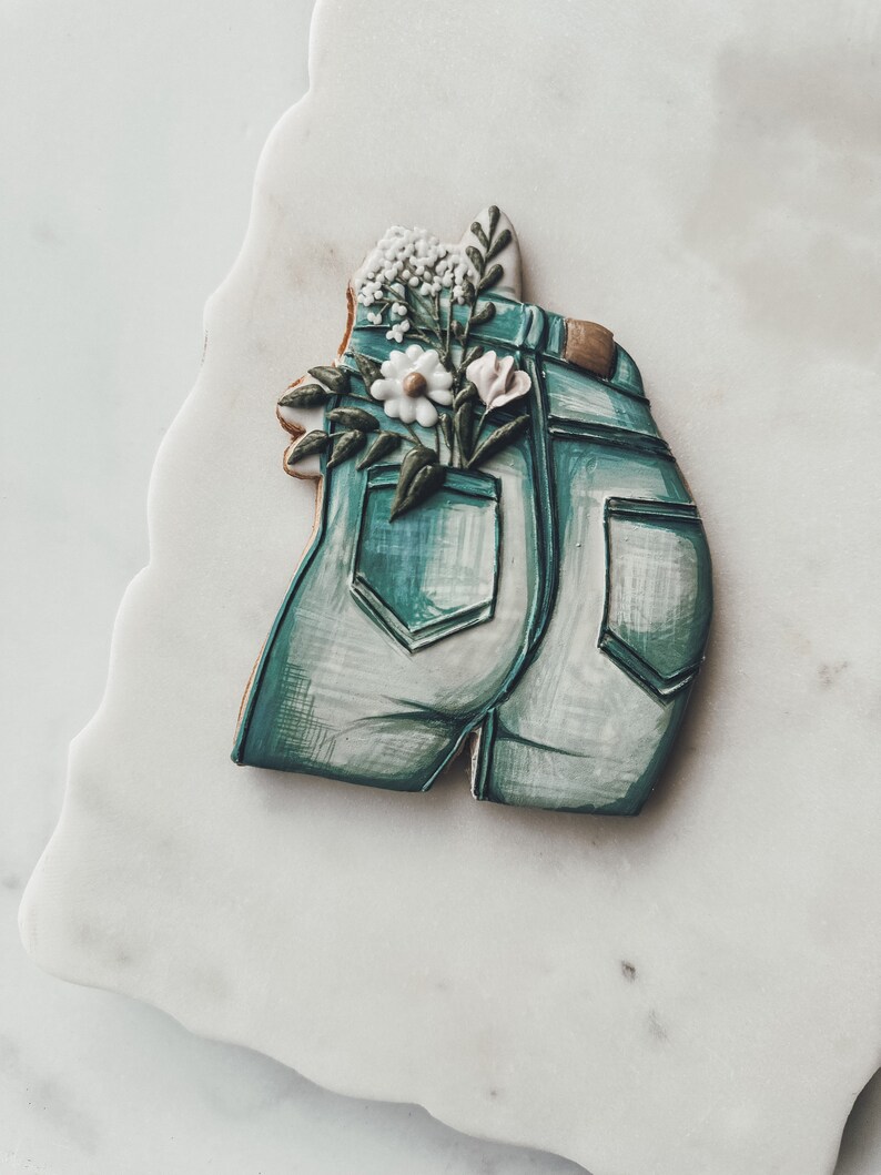 K&ouml;nnte beinhalten: Ein dekorierter Zuckerkeks in Form von Jeansshorts mit einem Blumenarrangement in der Ges&auml;&szlig;tasche. Der Keks ist hellblau mit wei&szlig;en Schattierungen und braunen Akzenten. Die Blumen sind wei&szlig;, rosa und gr&uuml;n.
