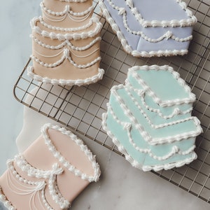 Puede incluir: Cuatro galletas de azúcar decoradas con forma de pasteles de varios pisos. Las galletas están decoradas con glaseado blanco y tienen una variedad de colores, incluyendo azul claro, verde claro, rosa claro y naranja claro.