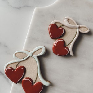 Puede incluir: Dos galletas de azúcar decoradas con corazones rojos y lazos blancos. Las galletas están sobre una superficie de mármol blanco.