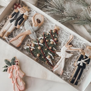 May include: A set of six Christmas-themed sugar cookies in a white box. The cookies are decorated with icing and feature a nutcracker, a Christmas tree, and ballerinas.