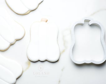 以下が含まれることがあります： 白い砂糖のクッキーがカボチャの形をしています。そのうちの1つのクッキーは、カボチャの形をしたクッキーカッターで切り抜かれています。クッキーは白い大理石の表面に置かれています。画像の右下隅には「LOLANO Cookie Cutters」というテキストが表示されています。