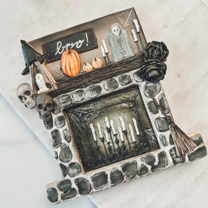 Pode incluir: Um biscoito de gengibre temático de Halloween decorado com uma lareira de pedra, um chapéu de bruxa, uma vassoura, uma lápida, abóboras e velas. O biscoito tem as palavras "boo!" escritas nele.