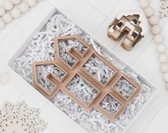 May include: A brown gingerbread house cookie cutter with a white background. The cutter is shaped like a house with windows and a door. The cutter is surrounded by white tissue paper.