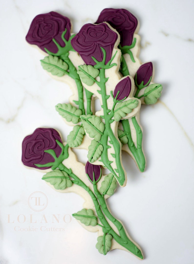 May include: A set of four sugar cookies decorated with purple roses and green leaves. The cookies are arranged in a bouquet shape. The cookies are made by Lolano Cookie Cutters.