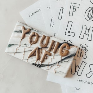 Könnte beinhalten: Eine Nahaufnahme eines dekorativen Schilds mit den Worten "YOUNG AF", die in goldenen Ballonbuchstaben geschrieben sind. Die Buchstaben sind an Schnüren befestigt, die herunterhängen. Das Schild hat einen marmorartigen Hintergrund mit schwarzen und weißen Adern.