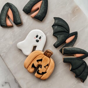 May include: Halloween-themed cookies, including a smiling ghost, a jack-o'-lantern, and bat wings. The cookies are decorated with black, orange, and peach-colored icing. The cookies are arranged on a marble surface.