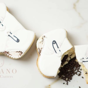 Peut inclure: Deux biscuits blancs en sucre décorés de glaçage pour ressembler à des bodys de bébé. Un body est entier, l'autre est cassé pour révéler des pépites de chocolat à l'intérieur. Les biscuits sont sur une surface en marbre blanc. Le texte "LOLANO Cookie Cutters" est visible sous les biscuits.