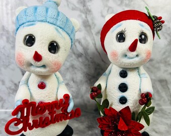May include: Two snowman figurines, one wearing a blue hat and holding a red sign that says "Merry Christmas", the other wearing a red hat and holding a red poinsettia.