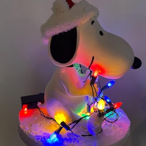 May include: A white ceramic figurine of Snoopy wearing a Santa hat and holding a string of colorful Christmas lights. The figurine is sitting on a white base with snow-like texture.
