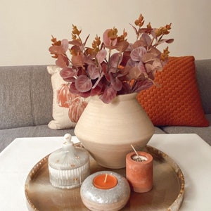May include: A round, light brown tray with gold accents, featuring a white ceramic vase with dried brown foliage, two small terracotta candle holders, and a white candle holder with a lit candle.