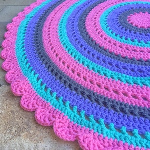 May include: A round crocheted rug with a scalloped edge. The rug is made with pink, teal, purple, and gray yarn.
