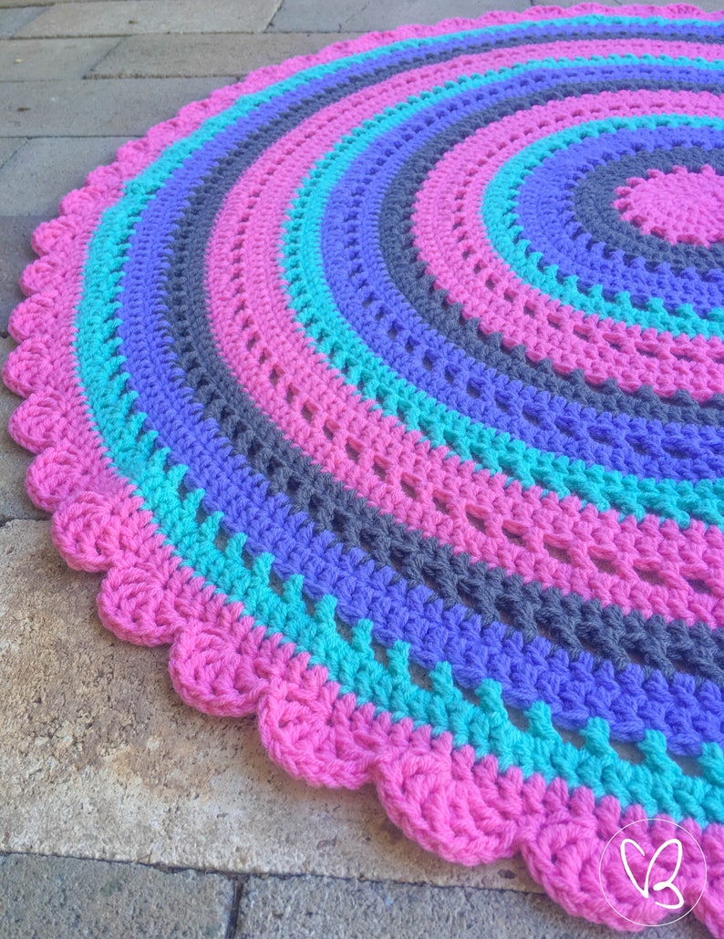 May include: A round crocheted rug with a scalloped edge. The rug is made with pink, teal, purple, and gray yarn.
