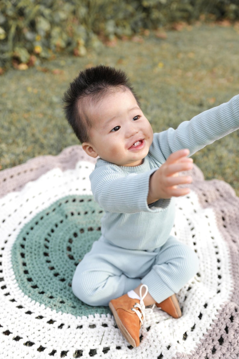 May include: A baby wearing a light blue long-sleeved outfit and brown shoes sits on a multi-colored crocheted rug. The baby is smiling and looking up.