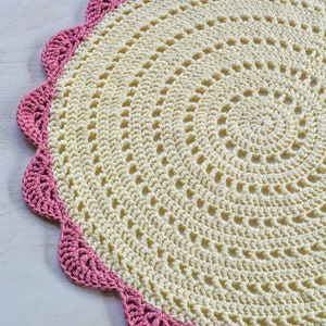 May include: A crocheted round doily with a scalloped edge. The doily is cream colored with a pink border.
