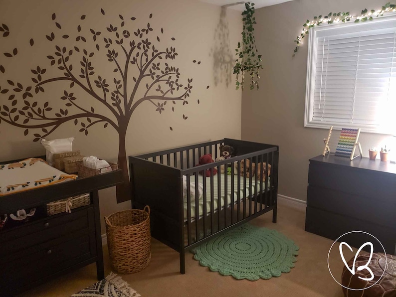 May include: A nursery room with a crib, a changing table, and a brown wicker basket. The walls are painted a light beige color and feature a brown tree decal. The crib has a white mattress and a green and white blanket. The changing table has a black changing pad and a white and yellow changing mat. The wicker basket is empty.