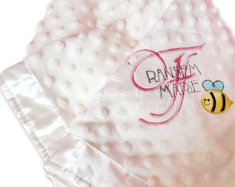 May include: White baby blanket with a satin border and pink embroidered lettering that says "Ransom Marie" with a bee design.