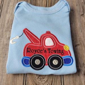 May include: A light blue baby onesie with an embroidered red tow truck with a hook, blue wheels, and the text "Royce's Towing" in black.