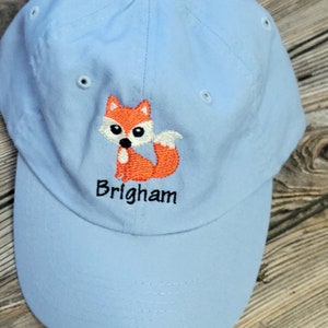 May include: A light blue baseball cap with an embroidered orange fox and the name "Brigham" in black.