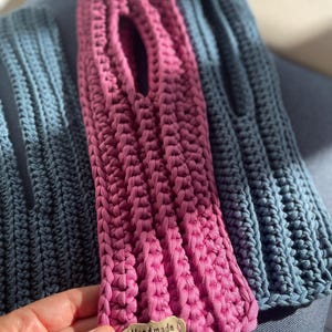 May include: A handmade, pink crochet bag with a rectangular shape and a large opening. The bag has a ribbed texture and a small tag that reads "Handmade with love". A blue crochet bag is visible in the background.