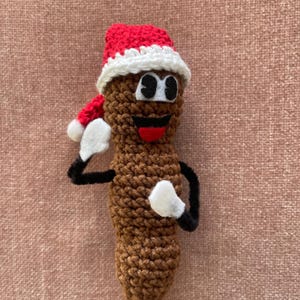 Puede incluir: Un emoji de caca marrón tejido a ganchillo con un gorro de Papá Noel rojo y blanco, brazos negros y guantes blancos. El emoji tiene una cara sonriente con ojos negros y una lengua roja. El artículo de novedad es una decoración navideña humorística.