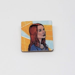 Könnte beinhalten: Eine quadratische Anstecknadel mit gelbem und blauem Hintergrund. In der Mitte der Anstecknadel befindet sich eine Comic-Illustration einer Frau mit roten Haaren, die eine blaue Jacke trägt.