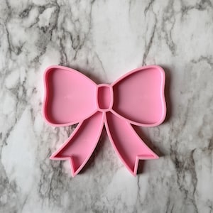 May include: A pink bow-shaped cookie cutter. The cutter is made of plastic and has a detailed design, including the bow's loops and tails. The cutter is designed for making bow-shaped cookies or other treats.