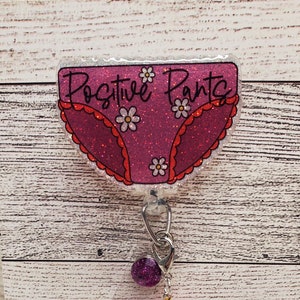 May include: A pink glitter badge holder with the text "Positive Pants" and a pink and white floral design. The badge holder has a clear plastic chain with a pink glitter charm and two pink beads.
