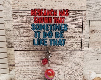 May include: A wooden sign with the text "Research has shown that sometimes it do be like that". The sign has a red and blue glitter finish. A charm with red and blue beads hangs from the sign.