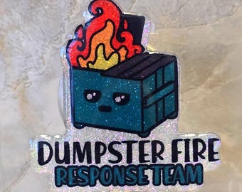 May include: A glittery badge reel featuring a teal dumpster on fire with the text "DUMPSTER FIRE RESPONSE TEAM". The reel has a clear plastic clip and a beaded charm with green, red, and orange beads.
