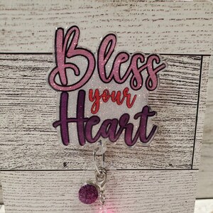 May include: A badge reel with the phrase "Bless your Heart" in pink and purple glitter lettering. The reel has a purple glitter accent bead, a pink bead, and a purple bead with white hearts.