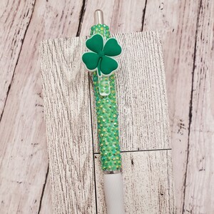 May include: A green pen with a white grip and a four-leaf clover charm. The pen is embellished with sparkling green rhinestones. The pen is resting on a white wooden background.