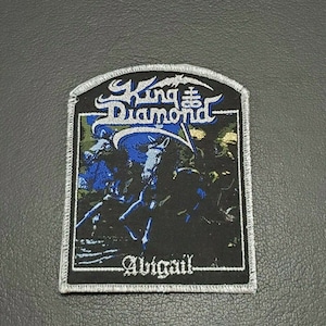 May include: A black and white embroidered patch featuring the band name "King Diamond" and the album title "Abigail" in white lettering. The patch has a dark background with a blue and green image of a figure with a sword.