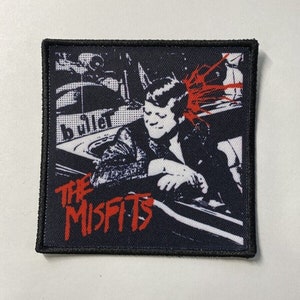 Könnte beinhalten: Schwarzer und weißer gestickter Patch mit einem roten und weißen Band. Der Patch zeigt einen Mann mit Irokesenschnitt und Sonnenbrille, der in einem Auto sitzt. Der Text "THE MISFITS" ist in roten Buchstaben unter dem Bild geschrieben.