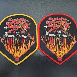 Puede incluir: Dos parches de la banda King Diamond, uno con borde amarillo y otro con borde rojo. Ambos parches presentan una imagen en blanco y negro de King Diamond con un traje rojo y negro, con llamas y el nombre de la banda en una fuente estilizada.