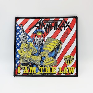 Könnte beinhalten: Ein schwarzer und roter gestickter Patch mit dem Bandnamen "Anthrax" und dem Text "I Am The Law" in Gelb. Der Patch zeigt eine Comic-Illustration eines Charakters mit Helm und Rüstung, der eine Waffe hält.