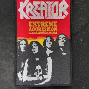 Puede incluir: Un parche en blanco y negro con el nombre de la banda "Kreator" en letras plateadas y las palabras "Extreme Aggression" en letras amarillas. El parche muestra a cuatro miembros de la banda en blanco y negro.