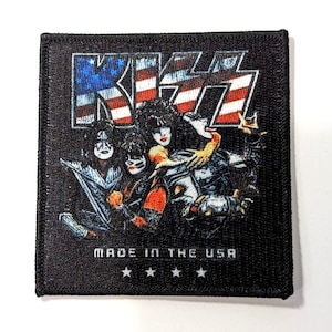 May include: A black fabric patch with a printed image of the rock band KISS. The band members are shown in front of an American flag with the band's name in large white letters. The text "MADE IN THE USA" is printed below the image with four stars.