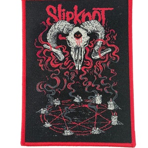 Puede incluir: Parche bordado en negro y rojo con el nombre de la banda "Slipknot" y una calavera con cuernos y un ojo en el centro. La calavera está rodeada de llamas rojas y un pentagrama rojo con velas.
